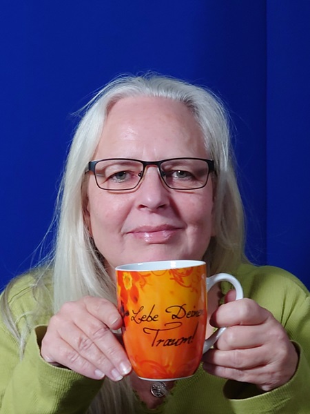Alexandra vor einem blauen Hintergrund mit einer orangefarbenen Tasse mit der Aufschrift "Lebe deinen Traum"