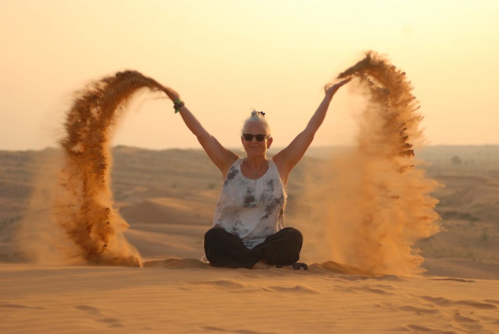 Alexandra sitzt im Schneidersitz in der Wüste auf dem Sand und wirft diesen mit beiden Händen in die Luft