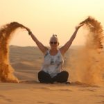 Alexandra sitzt im Schneidersitz in der Wüste auf dem Sand und wirft diesen mit beiden Händen in die Luft