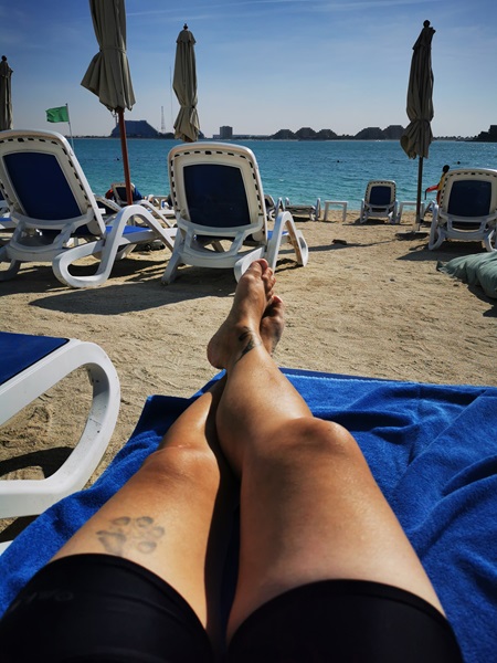 Alexandra liegt auf einer Liege am Strand. Es sind nur die Beine und Füße zu sehen. Im Hintergrund ein paar Liegestühle vor dem Meer