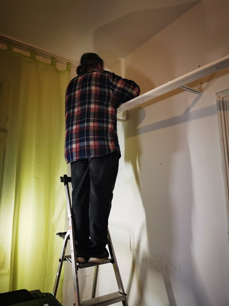 Ein Mann steht auf einer Leiter und schraubt eine Vorhangleiste an die Wand