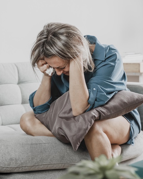 Frau sitzt weinend auf Sofa, hat ein Kissen auf dem Schoß, den Kopf in die Hände gestützt und hält ein Taschentuch in der Hand
