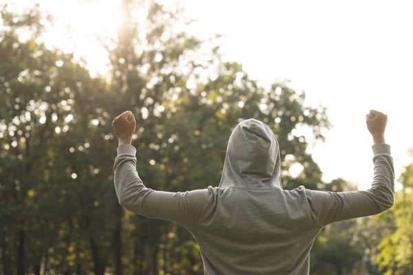 Person in der Rückansicht vor einem Wald stehend. Die Arme sind zu den Seiten hin noch oben gehalten und die Fäuste zu einer Siegerpose geballt.