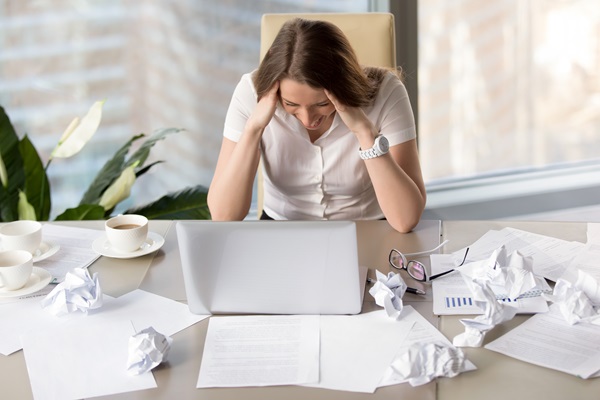 Frau sitzt schreiend am Schreibtisch und hat den Kopf in die Hände gestützt. Auf dem Tisch stehen eine Kaffeetasse und der Laptop. Drumherum liegen überall zusammengeknüddelte Papierblätter.