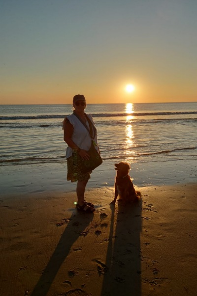 Eine Frau steht mit ihrem Hund am Sandstrand. Im Hintergrund ist ein schöner Sonnenuntergang über der ganz ruhigen Nordsee zu sehen.