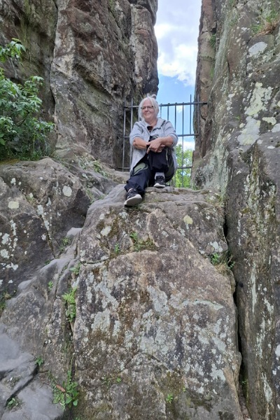 Alexandra sitzt, ein Knie umschlungen, auf einem hohen Felsen. Im Hintergrund ist ein rostiges Gitter zwischen zwei weiteren hohen Felswänden zu sehen. Und dahinter ein blauer Himmel mit kleinen weißen Wölkchen.