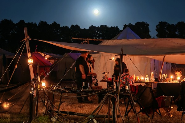 Ein mittelalterliches Lager bei Nacht mit gemütlicher Beleuchtung durch Kerzen, Fackeln, Laternen und Feuerschalen. Drei Personen, die aber durch die Entfernung zur Kamera, nur ein bisschen zu sehen sind, stehen unter einem gespannten Leinensegel. Dort steht auch eine lange Tafel, an der sicher 10 Personen Platz hätten. Auf der Tafel stehen auch nochmal ganz viele Kerzen und Öllampen. Im Hintergrund sieht man noch die schwarzen Umrisse von Bäumen und darüber, an dem tiefdunklen Himmel, sieht man den Vollmond. 