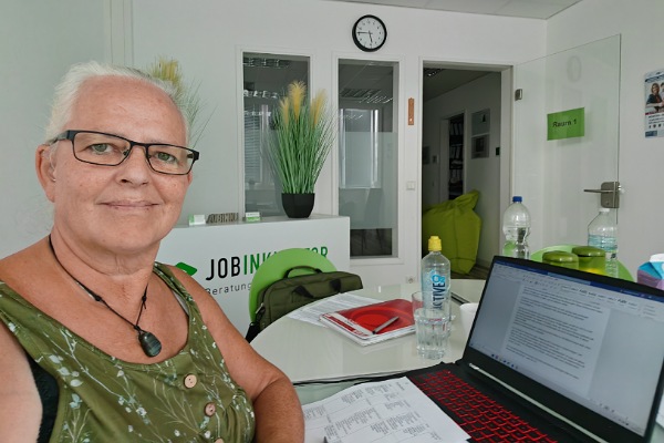 Alexandra sitzt an einem Schreibtisch in einem großen Büroraum. Sie trägt ein  schulterfreies, grünes Oberteil und lächelt in die Kamera. Auf dem Schreibtisch vor ihr stehen ein Laptop und Getränke. Auf der Tastatur des Laptops liegen ein Stapel Arbeitsblätter. Daneben liegen ein Collegeblock, ein Stift und weitere Arbeitsblätter. Im Hintergrund des Büros sieht man ein weißes Sideboard mit zwei Dekoschalen mit grünem Pampasgras, dahinter mehrere Glasfronten zum Flur hin und darüber, an der Wand, eine Uhr, die viertel vor sechs anzeigt. An der geöffneten Glastür hängt ein grünes Schild mit der Aufschrift "Raum 1" und im Flur erkennt man auf dem Boden einen großen grünen Sitzsack mit zwei kleineren Bildern, die an der Wand hängen.