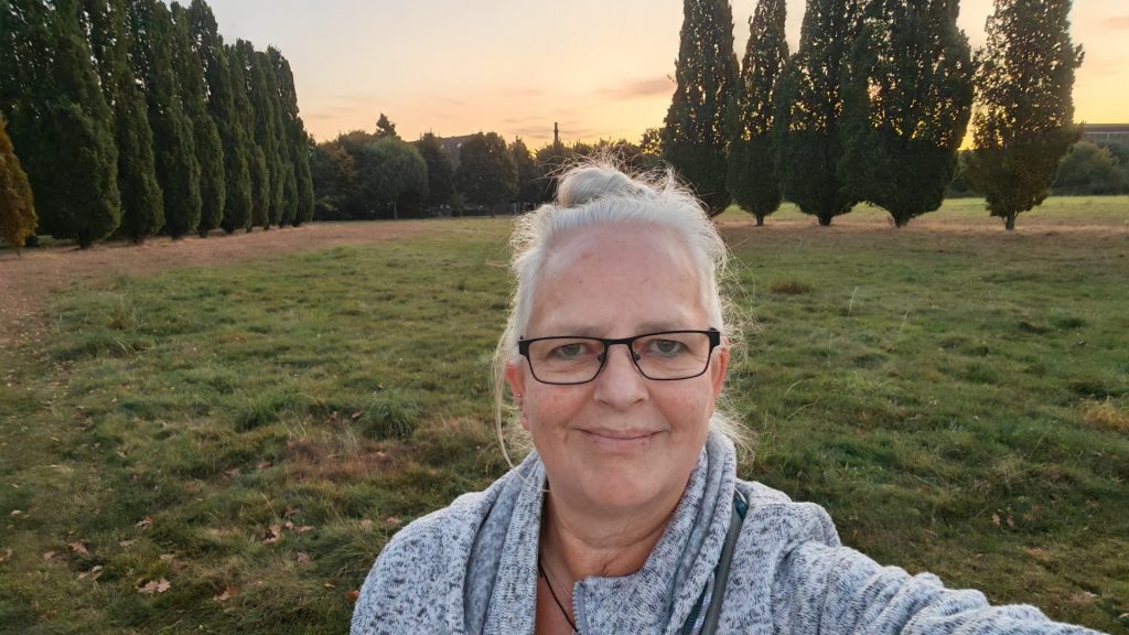 Alexandra steht auf einer grünen Wiese. Sie trägt eine weiß-grau-melierte Jacke und die weißen Haare am Hinterkopf zu einem lockeren Knoten gebunden. Sie schaut lächelnd in die Kamera. Im Hintergrund sind viele Bäume und ein blass-orange-farbener Himmel zu sehen. Durch das Sonnenuntergangslicht wirken die leicht angetrockneten Gräser am Rande der Wiese fast rosé-farben.