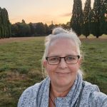 Alexandra steht auf einer grünen Wiese. Sie trägt eine weiß-grau-melierte Jacke und die weißen Haare am Hinterkopf zu einem lockeren Knoten gebunden. Sie schaut lächelnd in die Kamera. Im Hintergrund sind viele Bäume und ein blass-orange-farbener Himmel zu sehen. Durch das Sonnenuntergangslicht wirken die leicht angetrockneten Gräser am Rande der Wiese fast rosé-farben.