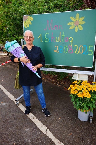 Alexandra steht auf einem Schulhof vor einer Schultafel auf der mit bunten Farben "Mein 1. Schultag 28.08.2025" geschrieben, und zwei Sonnenblumen gemalt, sind. Alexandra trägt eine blaue Jeans, schwarze Sneaker und ein schwarzes längeres Hemd. In der Hand hält sie eine große Schultüte, die fliederfarbene Schuppen aus Stoff hat, die untere Spitze und er obere Rand sind in türkis. Die Schultüte soll die Schwanzflosse einer Meerjungfrau darstellen. Vor der Tafel steht seitlich noch ein Blumentopf mit einer Pflanze mit grünen Blättern und gelben Blüten, die Sonnenblumen sehr ähnlich sind. 