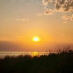 Das Titelbild zum Blogartikel "Was ist... Die Polyvagal-Therapie: Ein orangefarbener Sonnenuntergang über dem Meer. Im Vordergrund sind im Schattenbereich Dünen zu erkennen