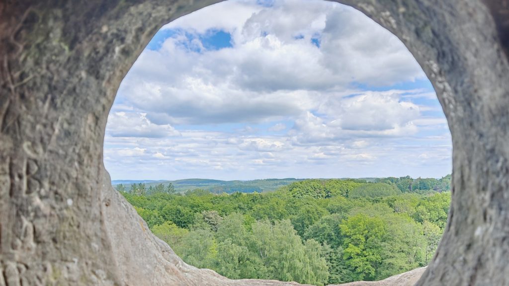 Der Blick durch ein Loch in einem dicken Mauerwerk. Am Horizont ist blauer Himmel mit ein paar weißen Wolken und die grünen Baumwipfel eines Waldes zu erkennen.