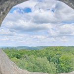 Der Blick durch ein Loch in einem dicken Mauerwerk. Am Horizont ist blauer Himmel mit ein paar weißen Wolken und die grünen Baumwipfel eines Waldes zu erkennen.