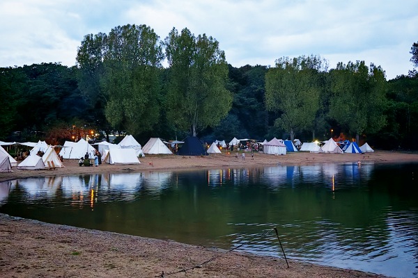 Im Vordergrund sieht man einen Sandstrand und einen großen See. Am gegenüberliegenden Ufer stehen, vor einem Wald, ganz viele verschiedene mittelalterliche Zelte. Die meisten mit weißen Tüchern, aber auch ein schwarzes und ein paar blau-weiße Zelte sind zu erkennen. In der Spiegelung im Wasser kann man auch sehr gut die teilweise Beleuchtung von Fackeln und Laternen erkennen.