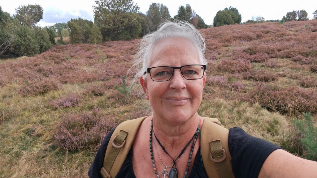 Alexandra steht mitten auf einem Feld mit langsam verblühendem rosa Heidekraut. Im Hintergrund sieht man viele Heidekrautpflanzen und am Horizont eine Linie mit verschiedenen grünen Bäumen, die das Feld abgrenzen. Alexandra trägt ihre schwarze Brille, mehrere ketten um den Hals, ein schwarzes T-Shirt und eine beigefarbenen Rucksack auf dem Rücken, von dem aber nur die Träger zu sehen sind ihre weißen Haare hat sie am Hinterkopf zusammen gebunden, durch den Wind werden aber viele einzelne Strähnen wild über den Kopf zu ihrer rechten Seite gepustet. Sie lächelt I'm die Kamera.