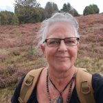 Alexandra steht mitten auf einem Feld mit langsam verblühendem rosa Heidekraut. Im Hintergrund sieht man viele Heidekrautpflanzen und am Horizont eine Linie mit verschiedenen grünen Bäumen, die das Feld abgrenzen. Alexandra trägt ihre schwarze Brille, mehrere ketten um den Hals, ein schwarzes T-Shirt und eine beigefarbenen Rucksack auf dem Rücken, von dem aber nur die Träger zu sehen sind ihre weißen Haare hat sie am Hinterkopf zusammen gebunden, durch den Wind werden aber viele einzelne Strähnen wild über den Kopf zu ihrer rechten Seite gepustet. Sie lächelt I'm die Kamera.