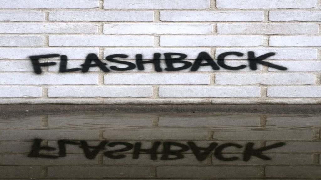 eine hellgraue Backsteinmauer, leicht verwittert, darauf das Graffiti FLASHBACK in schwarzen Spray-Buchstaben in Großschrift, etwas unregelmäßig. Im Vordergrund: eine Pfütze direkt vor der Mauer, leicht gewellte Wasseroberfläche, in der sich sowohl die Wand, als auch die gesamte Schrift spiegeln