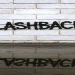 eine hellgraue Backsteinmauer, leicht verwittert, darauf das Graffiti FLASHBACK in schwarzen Spray-Buchstaben in Großschrift, etwas unregelmäßig. Im Vordergrund: eine Pfütze direkt vor der Mauer, leicht gewellte Wasseroberfläche, in der sich sowohl die Wand, als auch die gesamte Schrift spiegeln