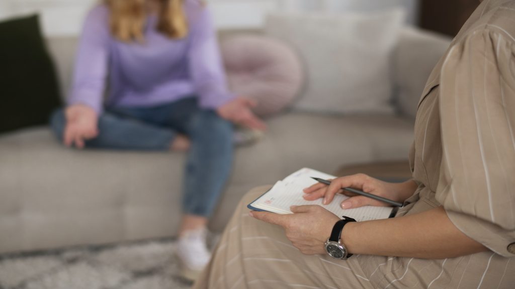 im Vordergrund sieht man einen Teil einer Frau in beigen Anziehsachen, die auf dem Schoß ein Notizbuch und einen Stift hält. Im Hintergrund ist, nur verschwommen zu erkennen, eine cremefarbene Couch, auf der eine andere Frau sitzt. Diese trägt eine blaue Jeans und einen Fliederfarbenen Pullover und hat blonde Haare