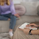 im Vordergrund sieht man einen Teil einer Frau in beigen Anziehsachen, die auf dem Schoß ein Notizbuch und einen Stift hält. Im Hintergrund ist, nur verschwommen zu erkennen, eine cremefarbene Couch, auf der eine andere Frau sitzt. Diese trägt eine blaue Jeans und einen Fliederfarbenen Pullover und hat blonde Haare