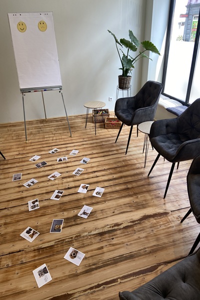 Workshop-Raum in der Praxis von Alexandra Winkens. Man sieht ein Flipchart an einer hellen Wand stehen. Rechts daneben steht auf dem Natur-Holzboden ein kleiner Hocker und zwei kleine Bastkörbchen. In der Ecke, zur großen Fensterfront hin, steht erhöht noch eine grüne Pflanze. Außerdem sieht man ein paar Stühle, die mit einem dunklen Stoff bezogen sind und in einem Halbkreis um das Flipchart herum aufgestellt sind. Auf den Holzdielen in der Mitte, liegen ein paar Karten verteilt.