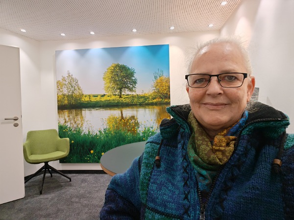 Alexandra Winkens sitzt in einer HNO Praxis. Ein hell erleuchteter Raum. An der Rückwand hängt ein sehr großes Bild mit einem Flusslauf, Bäumen am Ufer und grünen Wiesen. Davor steht ein grüner Stuhl. Alexandra sitzt im Vordergrund mit einer dicken, blau-türkis gemusterten Jacke.
