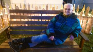 Titelbild zum Jahresrückblick 2025: Alexandra sitzt quer, mit den Füßen ausgestreckt, auf einer Holzbank. Hinter der Bank steht eine Art Holzzaun aus Paletten mit Gschnitzten Weihnachtsmotiven an den Spitzen. Der ganze Zaun ist mit einer warmweißen Lichterkette beleuchtet.