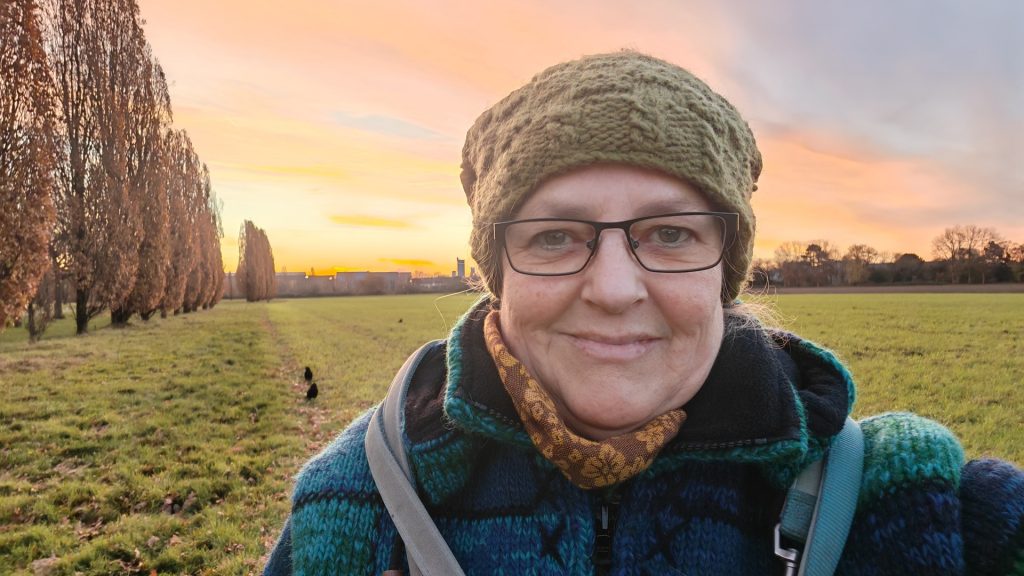 Titelbild zum Blogartikel Monatsrückblick November 2025. Das Bild zeigt Alexandra frühmorgens zum Sonnenaufgang, dick eingemummelt mit Mütze, Schal und dicker Jacke vor einem großen Feld. Im Hintergrund sieht man eine Krähe sitzen und der Himmel leuchtet in rosaroten bis gelben Farbtönen.
