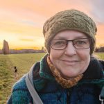 Titelbild zum Blogartikel Monatsrückblick November 2025. Das Bild zeigt Alexandra frühmorgens zum Sonnenaufgang, dick eingemummelt mit Mütze, Schal und dicker Jacke vor einem großen Feld. Im Hintergrund sieht man eine Krähe sitzen und der Himmel leuchtet in rosaroten bis gelben Farbtönen.