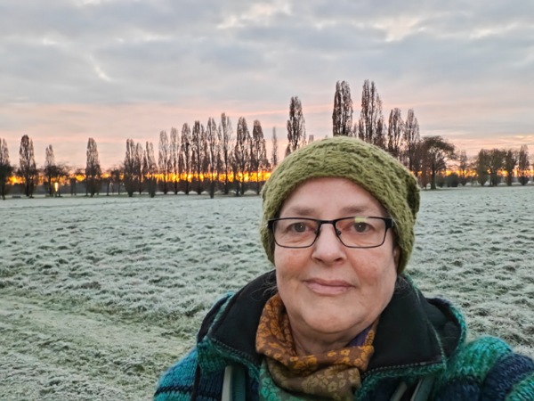 Ein Bild im Blogartikel Monatsrückblick Dezember 2025 von Alexandra Winkens zeigt Alexandra vor einem vereisten Feld. Ganz weit im Hintergrund sind noch Bäume zu sehen.