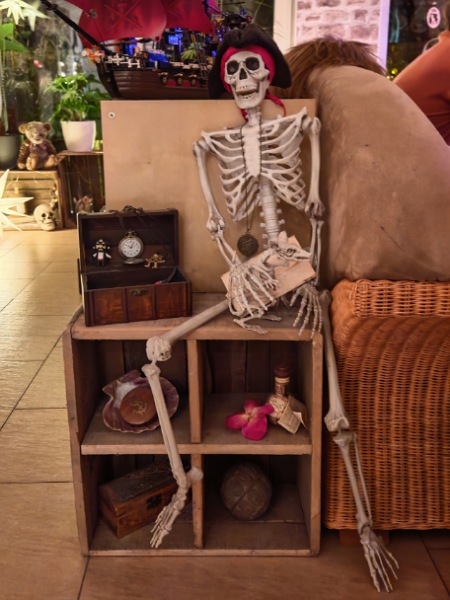 Bild zu einem Beitrag im Blogartikel Monatsrückblick Dezember 2025.
Ein Skelett sitzt auf einem kleinen Regal, an einen Schrank angelehnt. Auf dem Schrank steht ein Holzsegelschiff mit roten Segeln. Neben dem Skelett steht eine kleine, geöffnete Holztruhe. 