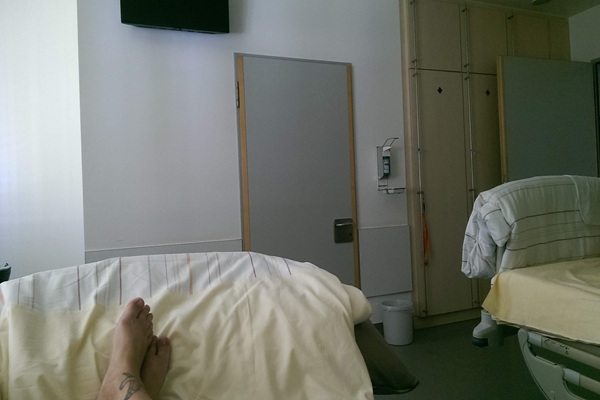 Alexandra Winkens liegt im Krankenhausbett in der Rheumatologie. Man sieht nur einen Fuß von ihr. An der weißen Wand gegenüber dem Bett sieht man eine Tür, daneben einen Desinfektionsspender und einen mehrtürigen Kleiderschrank.