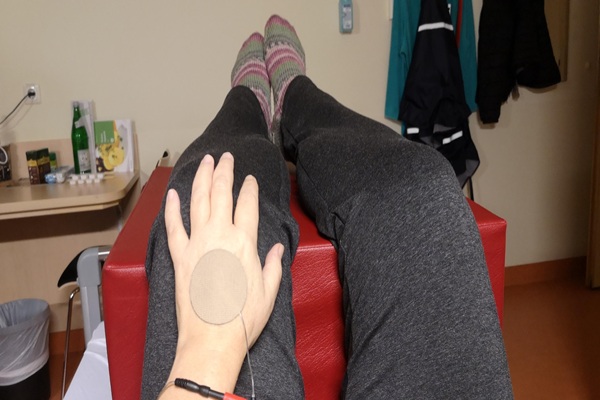 Alexandra Winkens in der Schmerzklinik. Sie liegt im Bett, die Beine auf einem großen, roten Würfel gelagert und hat ein Klebepad von einem Tens-Gerät auf der Hand kleben.