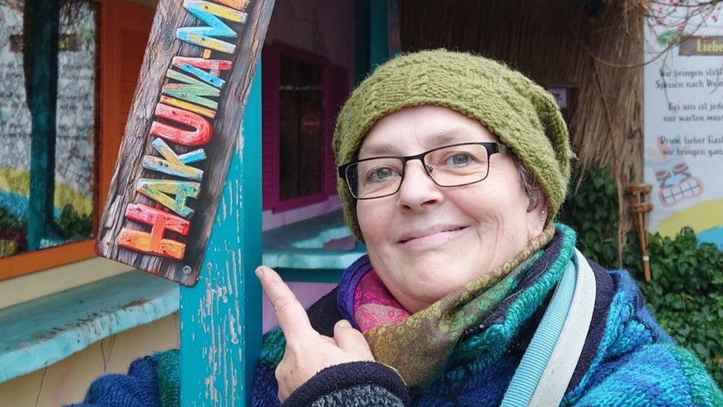 Titelbild zum Blogartikel "Funfacts" über mich" von Alexandra Winkens. Man sieht nur den Kopf von Alexandra. Sie trägt eine grüne Strickmütze, einen dicken bunten Schal und eine dicke, blaugrüngraue Strickjacke. Sie zeigt mit einem Finger auf ein buntes Schild auf dem steht: "HAKUNA-". Erahnen kann man, dass das zweite Wort Matata sein soll. Der obere Fotorand endet bei dem M.