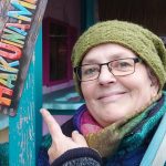 Titelbild zum Blogartikel "Funfacts" über mich" von Alexandra Winkens. Man sieht nur den Kopf von Alexandra. Sie trägt eine grüne Strickmütze, einen dicken bunten Schal und eine dicke, blaugrüngraue Strickjacke. Sie zeigt mit einem Finger auf ein buntes Schild auf dem steht: "HAKUNA-". Erahnen kann man, dass das zweite Wort Matata sein soll. Der obere Fotorand endet bei dem M.