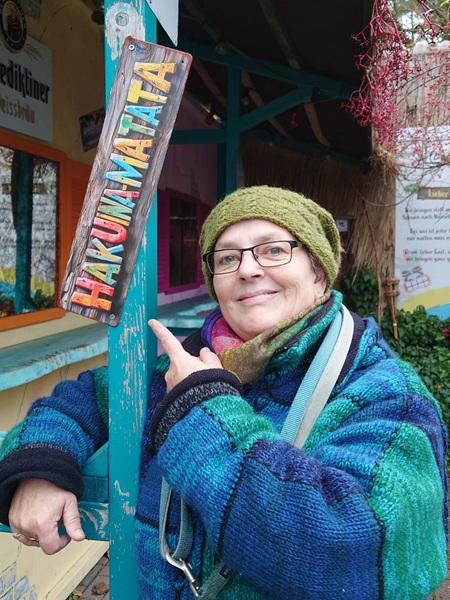 Noch einmal das Foto von Alexandra Winkens, wie sie auf das Schild mit der Aufschrift Hakuna-Matata zeigt. Dieses Mal im Ganzen, nicht abgeschnitten. Man sieht den Oberkörper von Alexandra. Sie trägt eine grüne Strickmütze, einen dicken bunten Schal und eine dicke, blaugrüngraue Strickjacke. Sie zeigt mit einem Finger auf ein buntes Schild auf dem steht: "HAKUNA-MATATA".