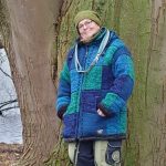 Titelbild zum Blogartikel Stress im Büro und Homeoffice - Der stille Stress des Sitzens. Alexandra Winkens steht an einen riesengroßen Baumstamm angelehnt. Im Hintergrund sieht man ein Gewässer. Der Stamm ist von Moos ganz grün. Alexandra trägt eine grüne dicke, Wollmütze in demselben Ton, wie das Moos, und eine dicke gestrickte Winterjacke mit großen blau und türkis-grünen Karos. Um den Hals hängt ihr locker eine blaue Hundeleine.