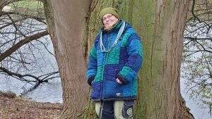 Titelbild zum Blogartikel Stress im Büro und Homeoffice - Der stille Stress des Sitzens. Alexandra Winkens steht an einen riesengroßen Baumstamm angelehnt. Im Hintergrund sieht man ein Gewässer. Der Stamm ist von Moos ganz grün. Alexandra trägt eine grüne dicke, Wollmütze in demselben Ton, wie das Moos, und eine dicke gestrickte Winterjacke mit großen blau und türkis-grünen Karos. Um den Hals hängt ihr locker eine blaue Hundeleine.