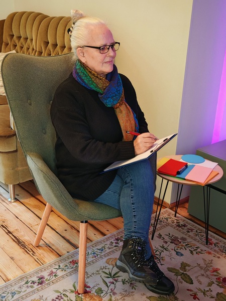 Alexandra sitzt, die Beine übereinandergeschlagen, ein Klemmbrett auf dem Schoß, im Therapie-Raum und schaut einen Klienten an.