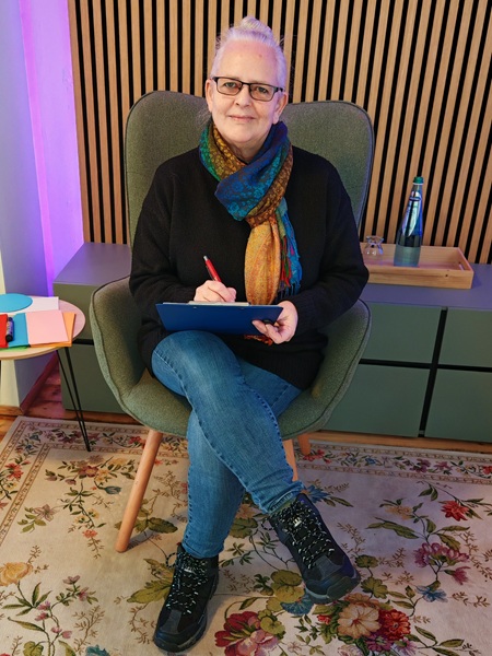 Alexandra sitzt auf einem Sessel, hat ein Klemmbrett im Schoß liegen und schaut lächelnd in die Kamera