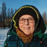 Beitragsbild zum Blogartikel "Monatsrückblick Januar2026". Alexandra steht, dick in Winterklamotten eingepackt auf einer mit Schnee bedeckten Wiese und lässt sich die Sonne ins Gesicht scheinen.